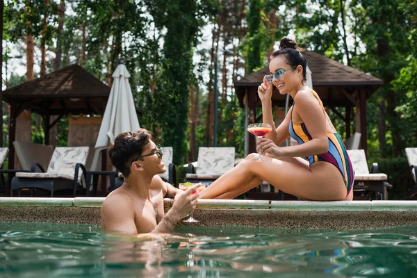 Позитивный мужчина держит стакан коктейля рядом с девушкой у бассейна — стоковое фото