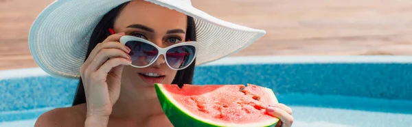 Молода жінка тримає сонцезахисні окуляри і кавун біля розмитого басейну, банер — стокове фото