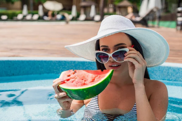 Frau mit Sonnenbrille hält Wassermelone in der Hand und blickt in Pool in Kamera — Stockfoto