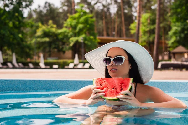 Молода жінка в сонячному капелюсі тримає шматок кавуна в басейні — стокове фото