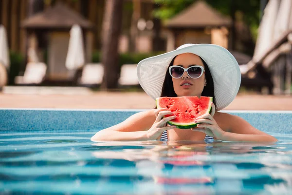 Brunette woman in sun hat holding piece of watermelon in swimming pool — Stock Photo