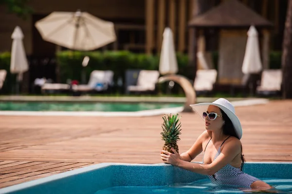 Young woman in swimwear holding pineapple in swimming pool — Stock Photo
