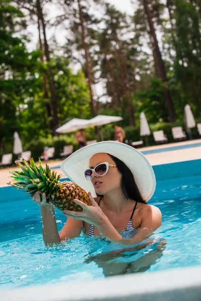 Mulher morena em óculos de sol olhando para abacaxi na piscina durante as férias — Fotografia de Stock