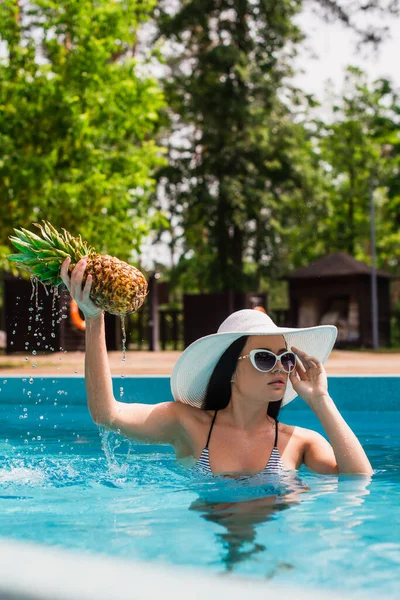 Young woman holding fresh pineapple with water drops and sunglasses in swimming pool — Stock Photo