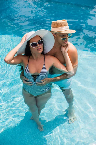 Vista de ángulo alto de hombre joven abrazando novia en sombrero de sol en la piscina - foto de stock