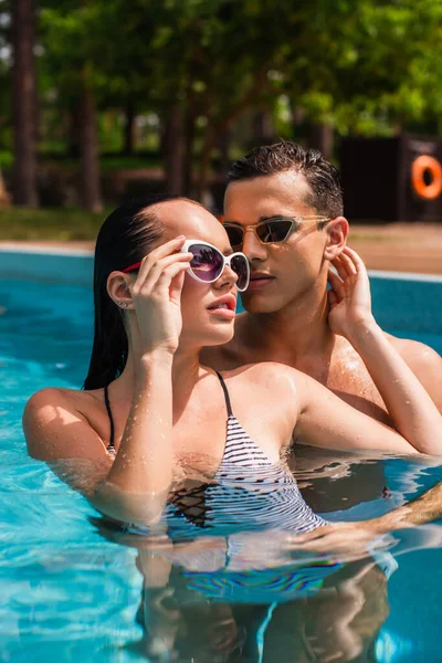 Jovem segurando óculos de sol e tocando namorado molhado na piscina — Fotografia de Stock