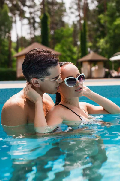 Sexy man kissing girlfriend in sunglasses in swimming pool — Stock Photo