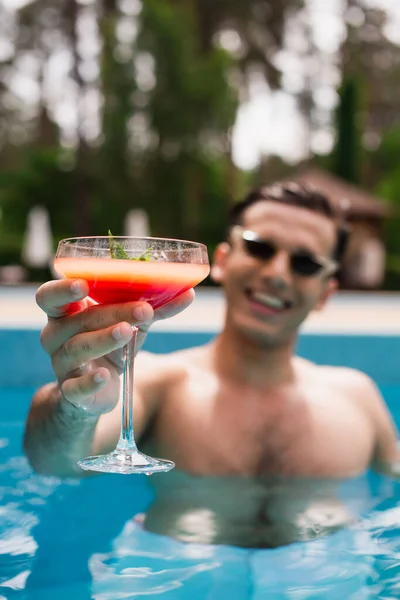 Copo de coquetel na mão de homem sorridente na piscina no fundo borrado — Fotografia de Stock