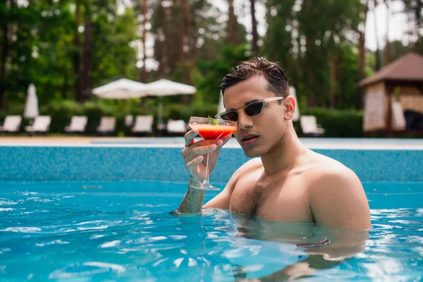 Молодий чоловік з мокрим волоссям тримає склянку коктейлю в басейні — стокове фото