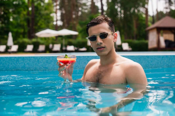 Young man with cocktail standing in swimming pool during summer — Stock Photo