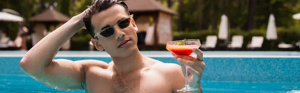 Jeune homme tenant un cocktail et ajustant les cheveux dans la piscine, bannière — Photo de stock