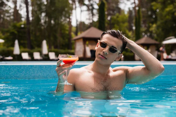 Jeune homme en lunettes de soleil ajustant les cheveux et tenant un cocktail dans la piscine — Photo de stock