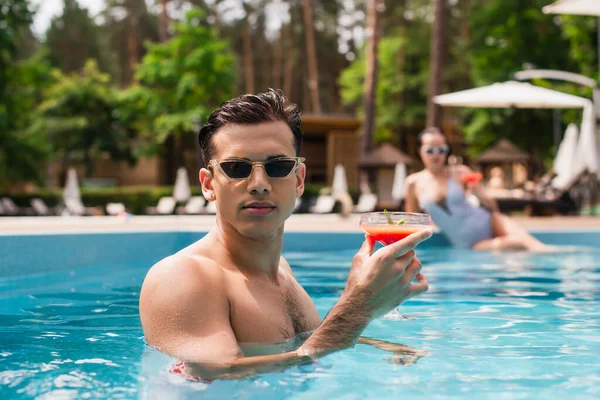 Young man holding cocktail near blurred girlfriend in swimming pool on blurred background — Stock Photo