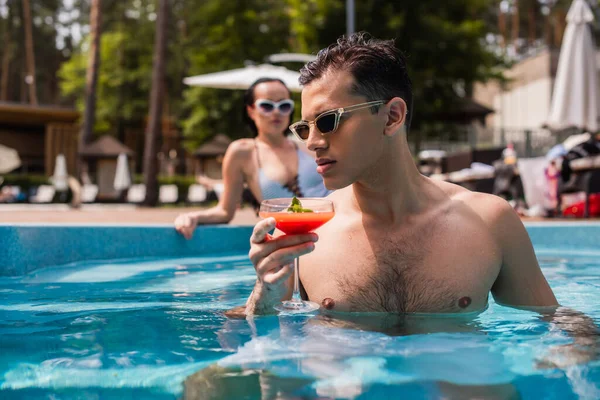 Uomo che tiene un bicchiere di cocktail in piscina vicino alla fidanzata offuscata sullo sfondo — Foto stock