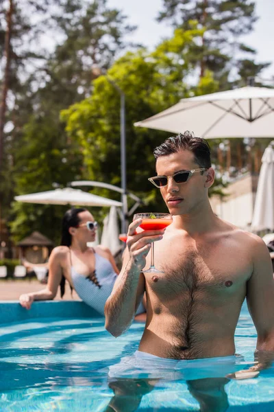 Homme avec lunettes de soleil et cocktail debout dans la piscine sur place — Photo de stock