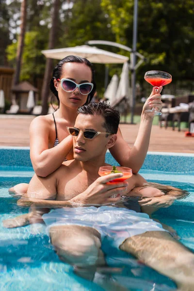 Mulher de óculos de sol segurando copo de coquetel perto de namorado na piscina — Fotografia de Stock
