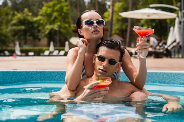 Mujer morena sosteniendo cóctel mientras se relaja cerca de novio en la piscina - foto de stock