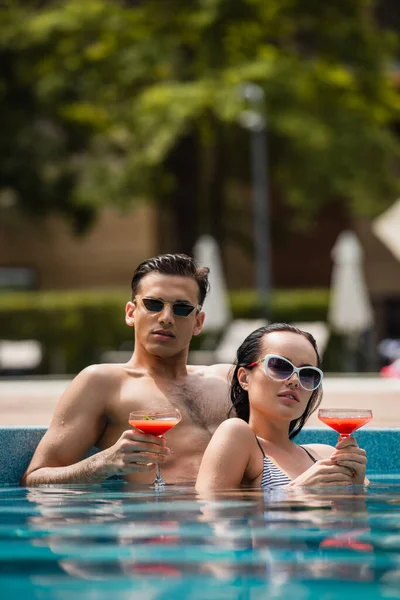 Nivel de superficie de la pareja joven en gafas de sol con cócteles en la piscina - foto de stock