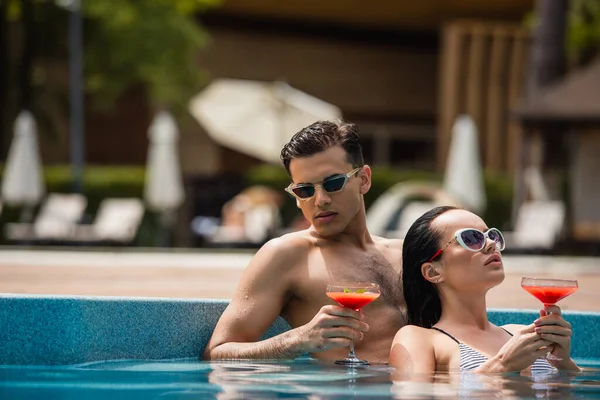 Homem segurando copo de coquetel perto de mulher morena na piscina — Fotografia de Stock