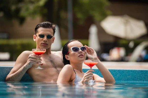 Nível de superfície da mulher ajustando óculos de sol perto do namorado com coquetel na piscina — Fotografia de Stock