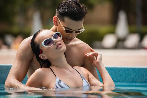 Mujer joven en traje de baño y gafas de sol tocando novio en la piscina - foto de stock