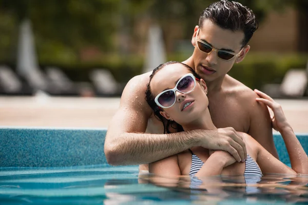 Homme avec les cheveux mouillés étreignant petite amie dans les lunettes de soleil dans la piscine — Photo de stock