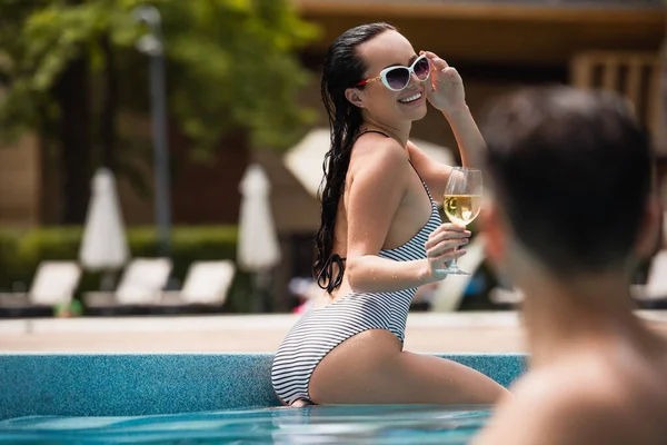 Mulher sorridente em maiô segurando copo de vinho perto de namorado na piscina em primeiro plano borrado — Fotografia de Stock
