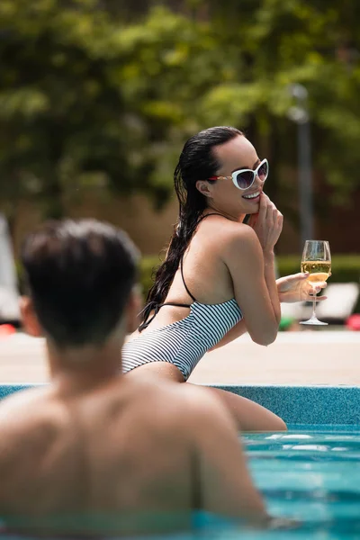 Mulher morena com copo de vinho perto do namorado em primeiro plano desfocado na piscina — Fotografia de Stock