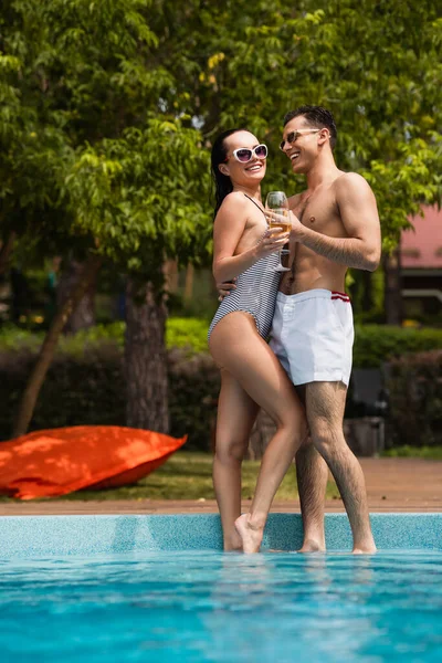 Pareja alegre en traje de baño abrazando y sosteniendo copas de vino cerca de la piscina - foto de stock