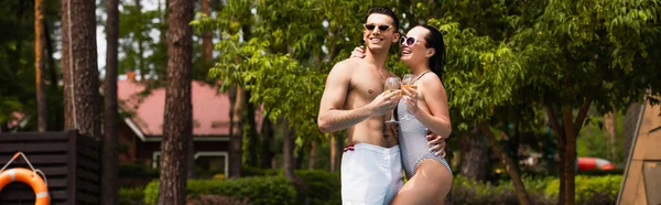 Mujer en traje de baño y gafas de sol abrazando novio con vino al aire libre, pancarta - foto de stock