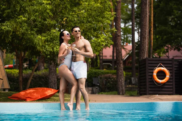 Smiling woman in swimwear holding wine and hugging boyfriend near pool on resort — Stock Photo