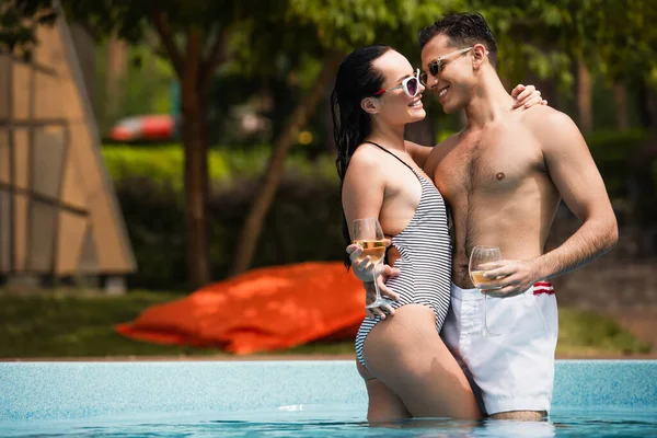 Pareja alegre abrazándose y sosteniendo copas de vino en la piscina - foto de stock