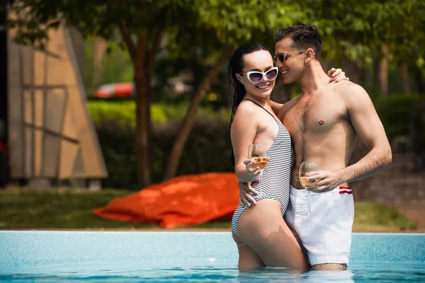 Sorrindo homem em óculos de sol e calções de banho segurando copo de vinho e abraçando namorada na piscina — Fotografia de Stock