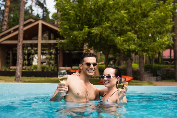 Cheerful couple in sunglasses holding glasses of wine in swimming pool outdoors — Stock Photo