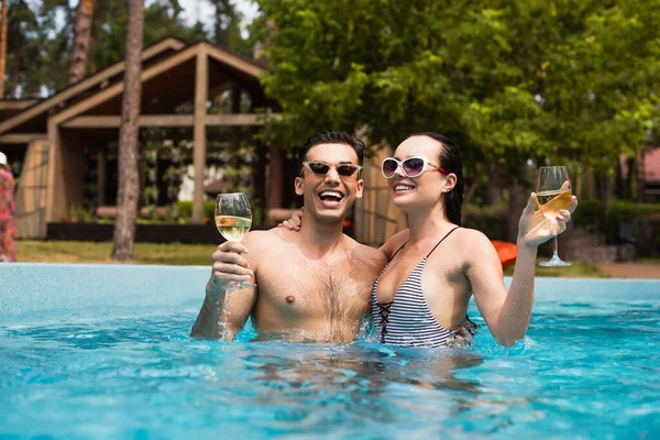 Couple joyeux dans des lunettes de soleil tenant des verres de vin dans la piscine — Photo de stock