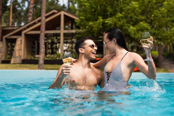 Smiling couple holding glasses of wine near splashes in swimming pool — Stock Photo