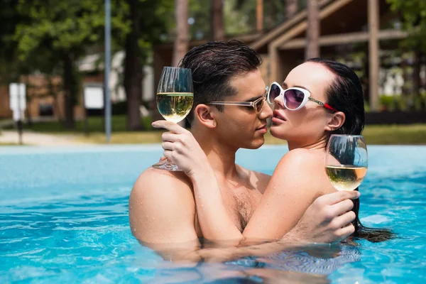 Couple in sunglasses holding wine and kissing in swimming pool — Stock Photo