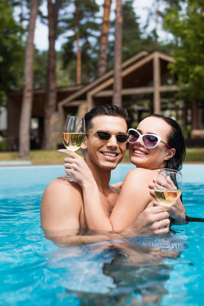 Молода пара в сонцезахисних окулярах посміхається на камеру, тримаючи біле вино в басейні — стокове фото