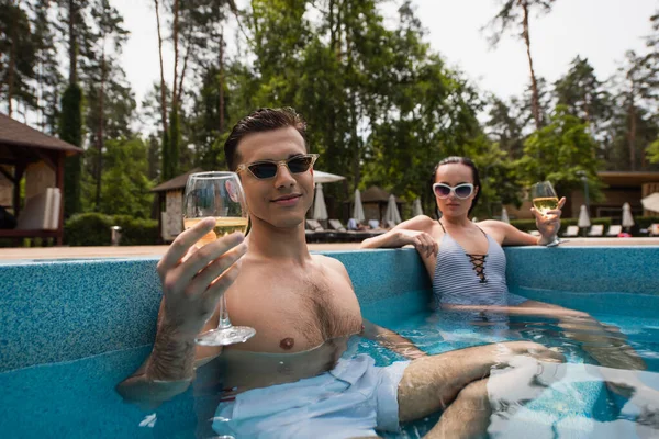 Homem segurando copo de vinho perto de namorada borrada na piscina — Fotografia de Stock