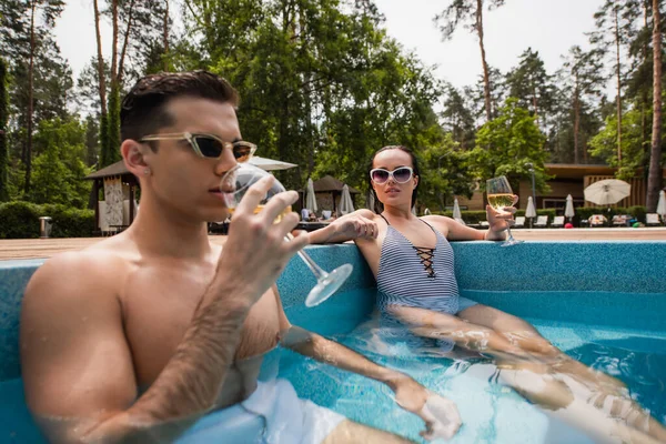 Молодая женщина в полосатых купальниках сидит рядом с парнем и пьет вино в бассейне. — стоковое фото