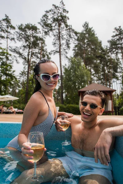 Sonriente hombre sentado en la piscina cerca de la novia sosteniendo borrosa copa de vino durante las vacaciones - foto de stock