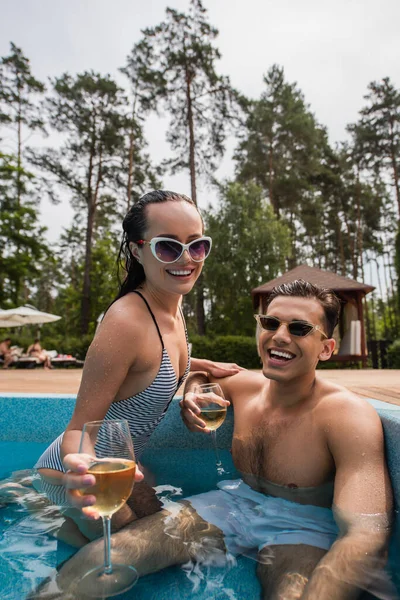 Mulher alegre com vinho borrado relaxante perto namorado molhado na piscina — Fotografia de Stock