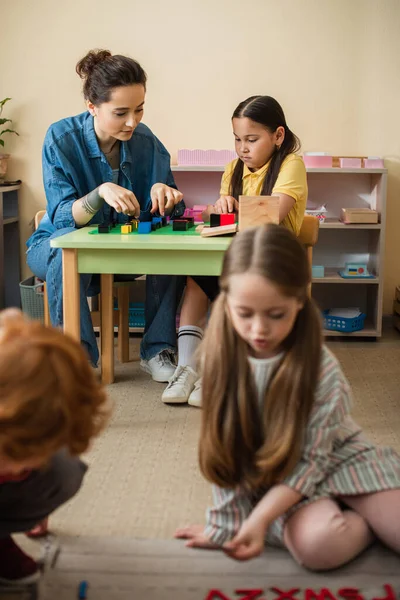 Menina asiática jogando cubos de madeira jogo com professor perto de crianças em primeiro plano borrado — Fotografia de Stock