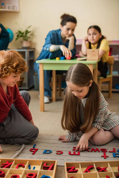 Kinder spielen mit Holzbuchstaben auf dem Boden neben verschwommenem Lehrer und asiatischem Mädchen — Stockfoto