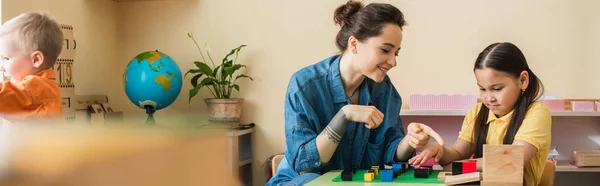 Montessori school teacher pointing at multicolored blocks while playing with asian girl, banner — Stock Photo