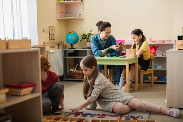 Kids on floor playing with wooden letters near teacher and girl with multicolored cubes on background — Stock Photo