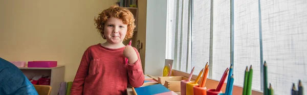 Lockiger, rothaariger Junge mit Stift in der Hand und lächelnd in die Kamera der Montessori-Schule, Banner — Stockfoto