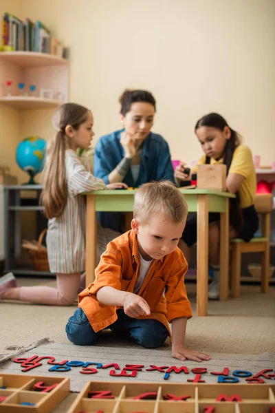 Boy on floor pointing at wooden letters near teacher and interracial kids on blurred background — Stock Photo