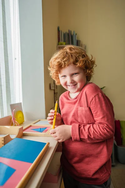 Alegre, ruiva menino olhando para a câmera enquanto segurando lápis de cor na escola montessori — Fotografia de Stock