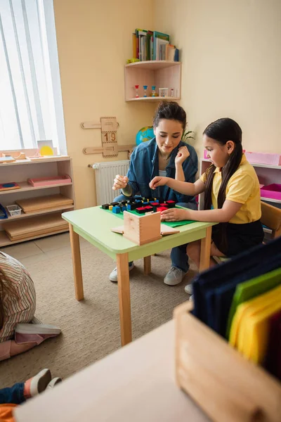 Menina asiática jogando cubos de madeira jogo com professor perto de crianças no chão em sala de aula — Fotografia de Stock